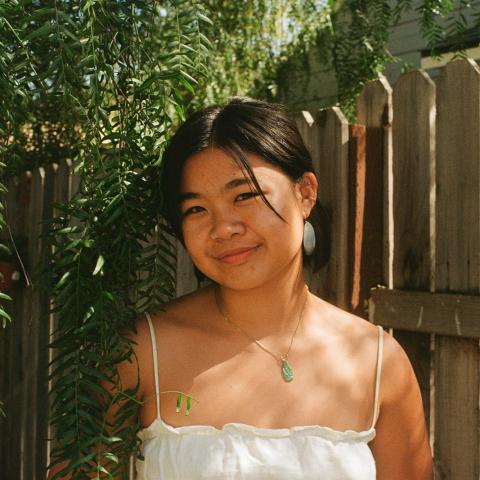 Natalie is standing in the middle of the frame, smiling at the camera. Natalie is framed by lush green foliage with a fence in the background.