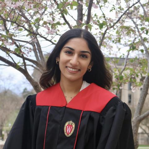 Anisha is standing in the middle of the frame, facing the camera and smiling. She is wearing black graduation regalia with red accents. Behind her is a tree with sparse foliage.